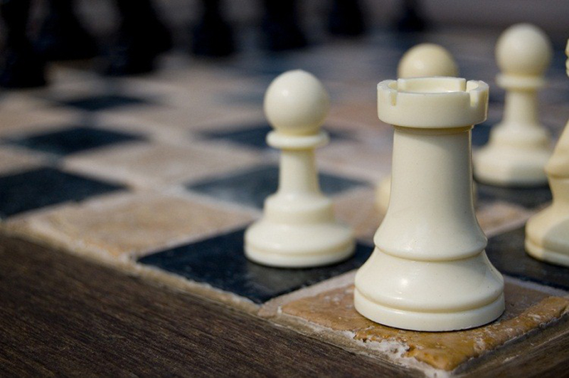 Closeup of white chess rook on tile game board with wood trim.